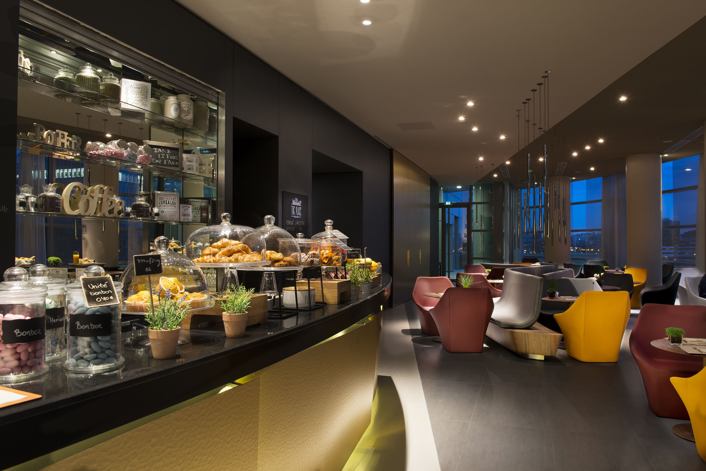 A modern cafe with a glass display case filled with pastries and a variety of coffee options. There are comfortable seating areas with colorful chairs and a view of the cityscape through large windows.