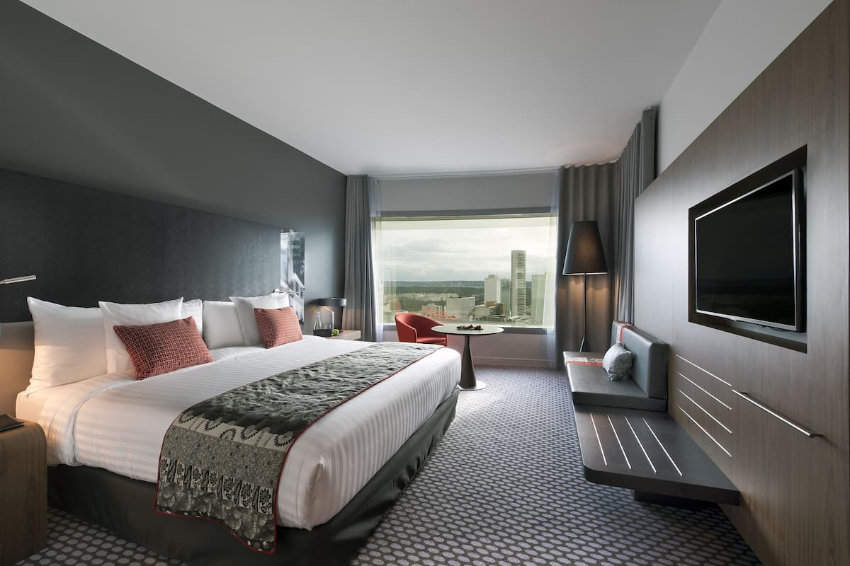A modern hotel room with a large bed, a seating area, a TV, and a city view.