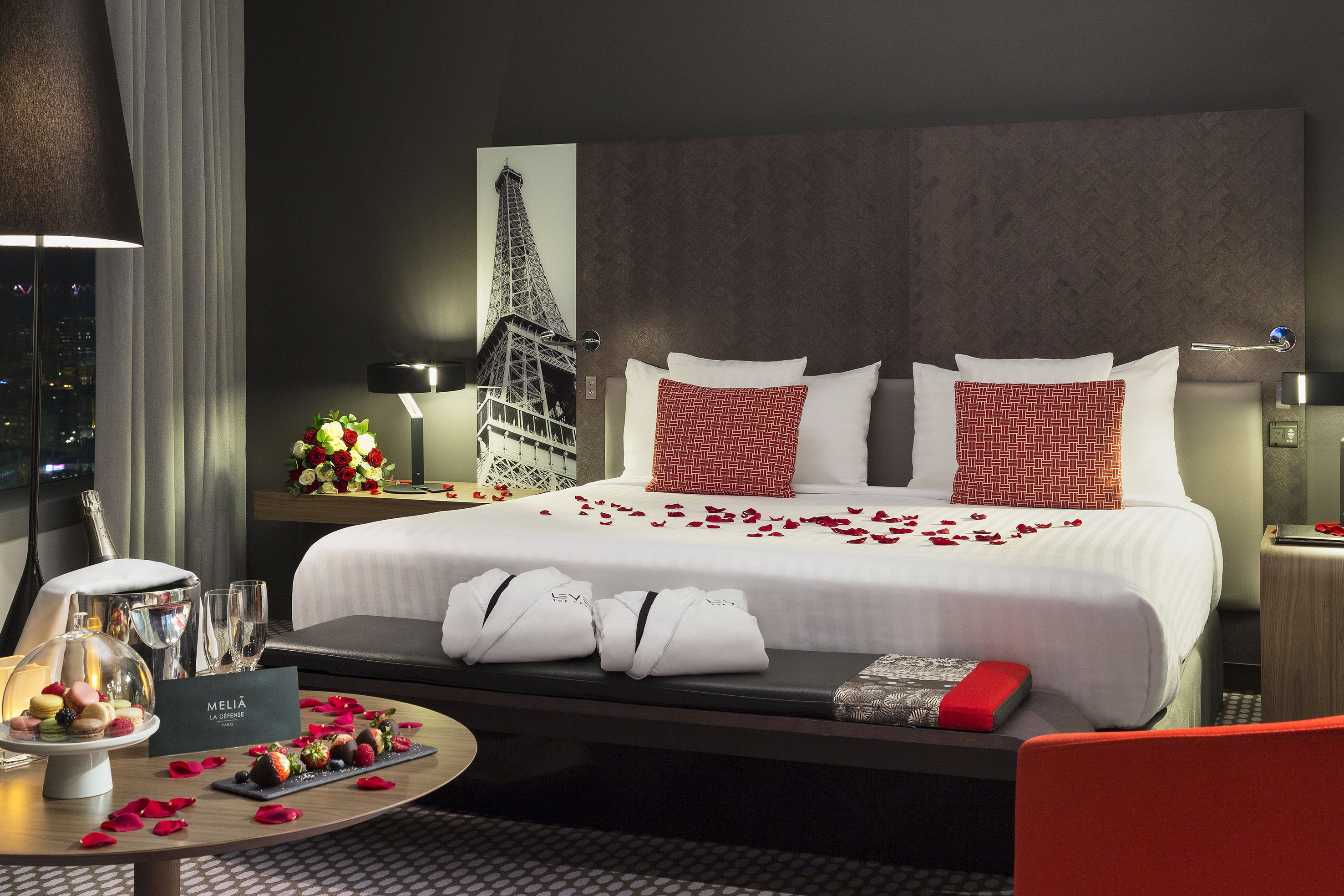 Habitación de hotel con cama, mesita de noche con lámpara, vista de la Torre Eiffel y paisaje urbano nocturno.