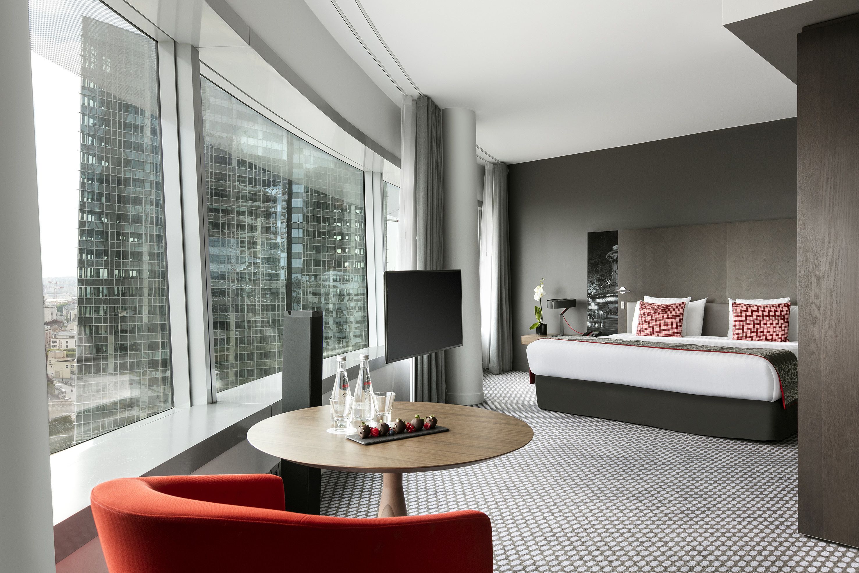 Una habitación de hotel moderna con un ventanal grande, una cama con ropa de cama roja y blanca, una mesa redonda con vasos y una bandeja de fresas, y una silla roja.