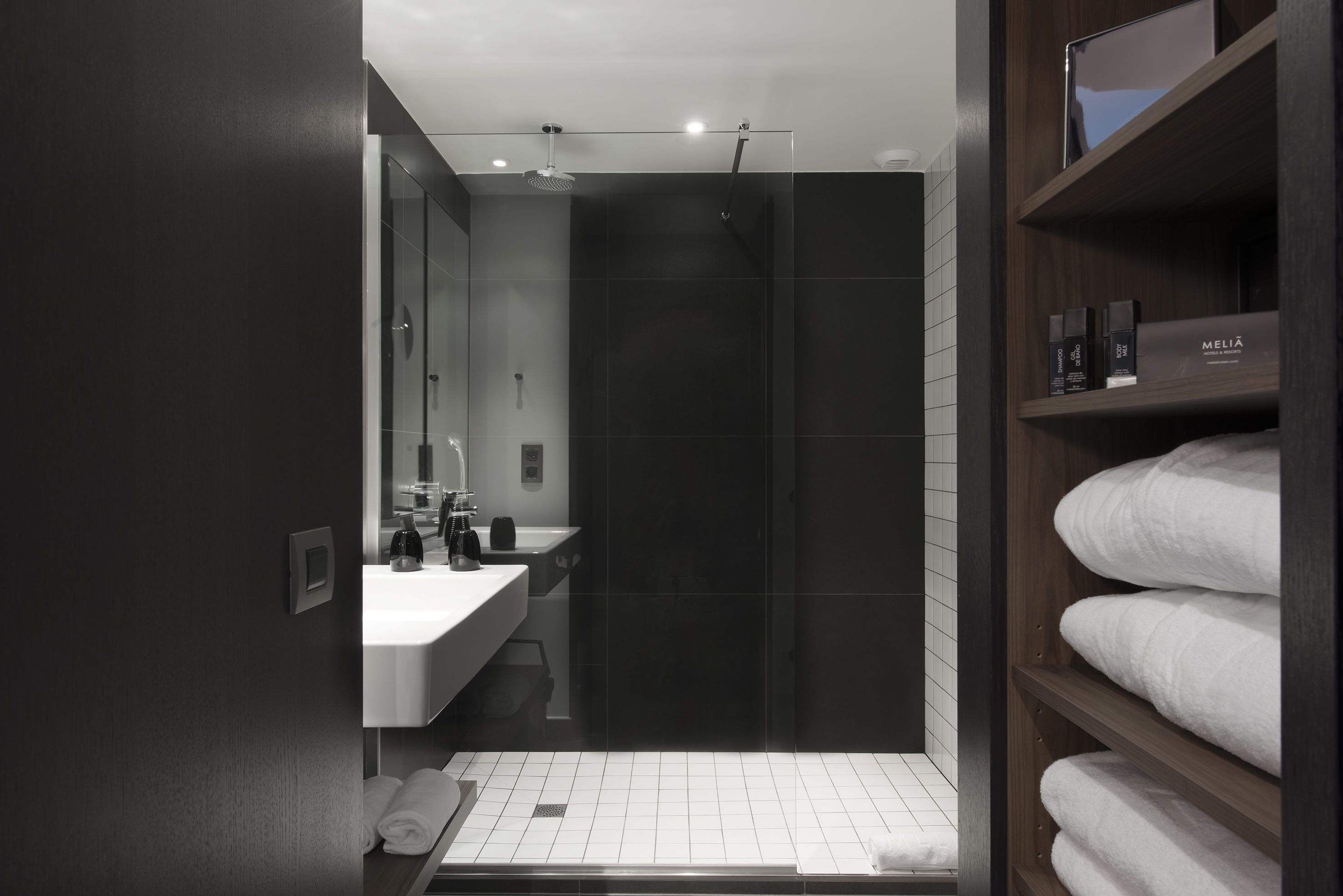 A modern bathroom with a glass shower enclosure, a white sink, and a wooden shelf with towels.