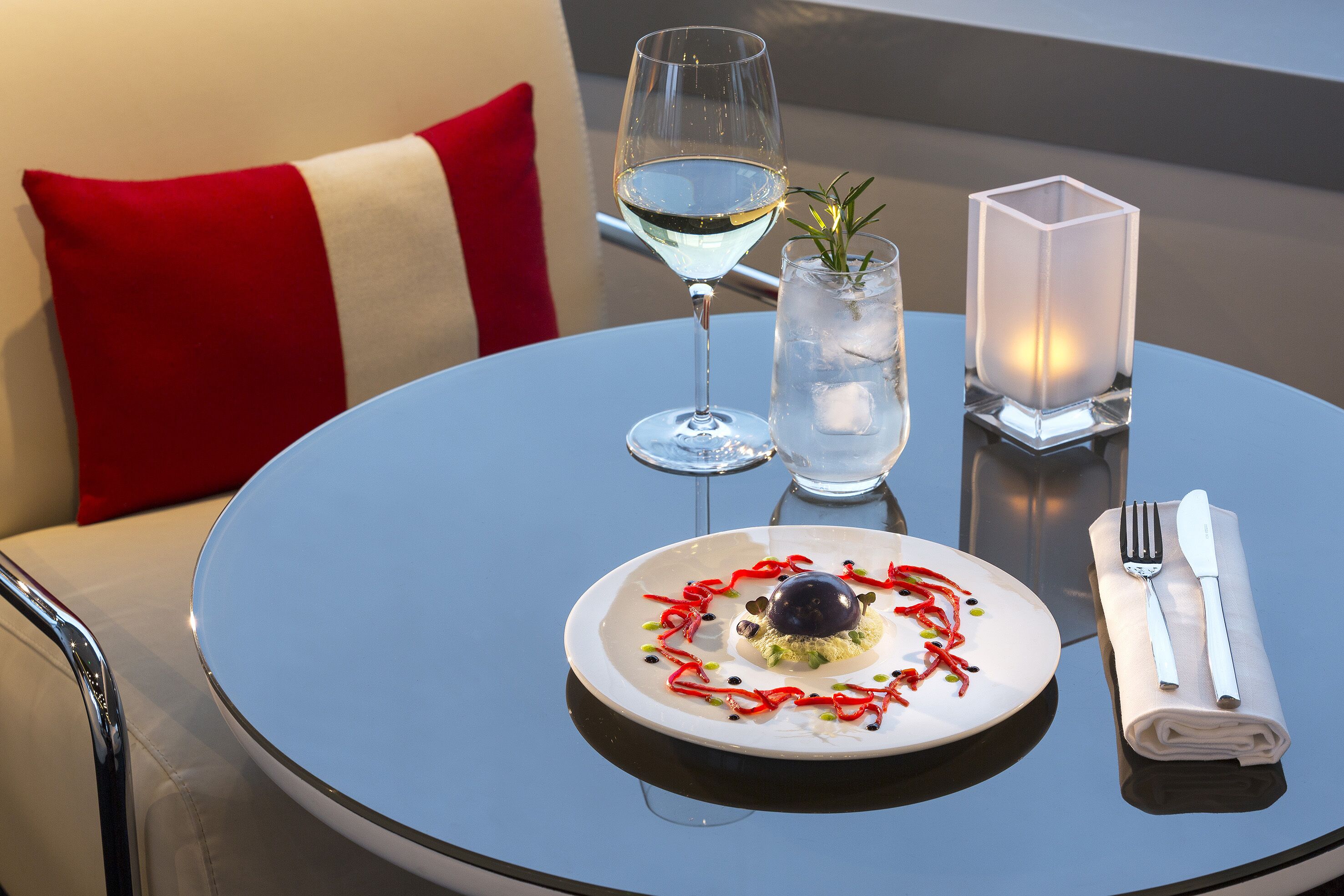 A dining table with a plate of food, a glass of white wine, a candle, and cutlery.