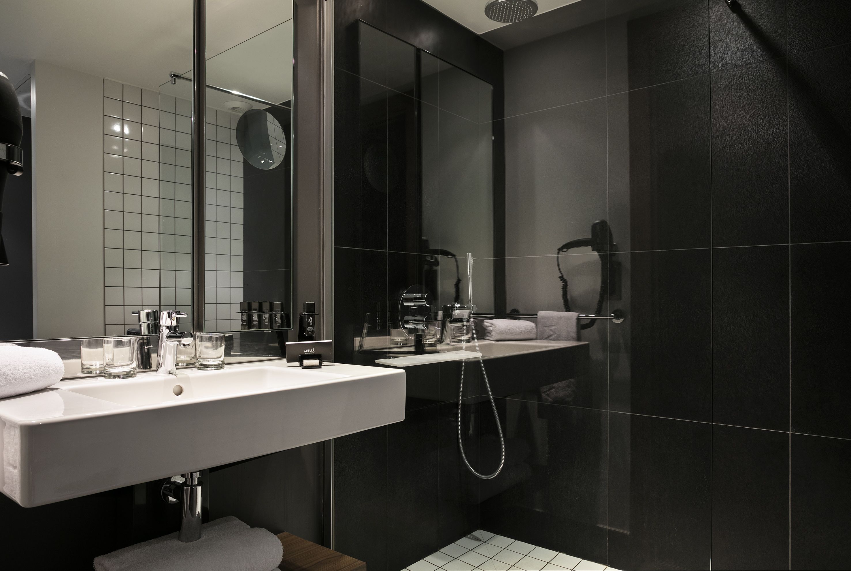 A modern bathroom with a white sink, a mirror, a shower, and dark tiled walls.