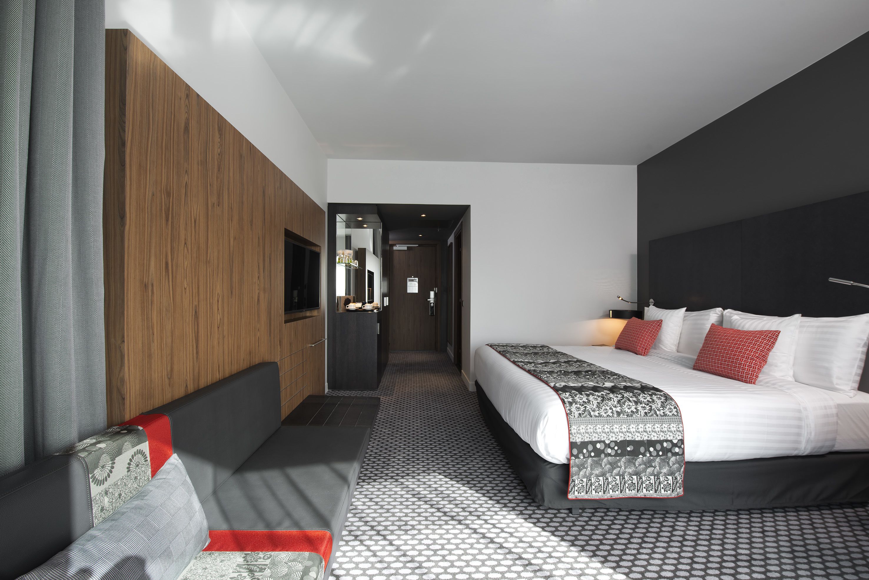 Une chambre d’hôtel moderne avec un grand lit, un canapé, un mur décoratif en bois et un couloir menant à la salle de bain.