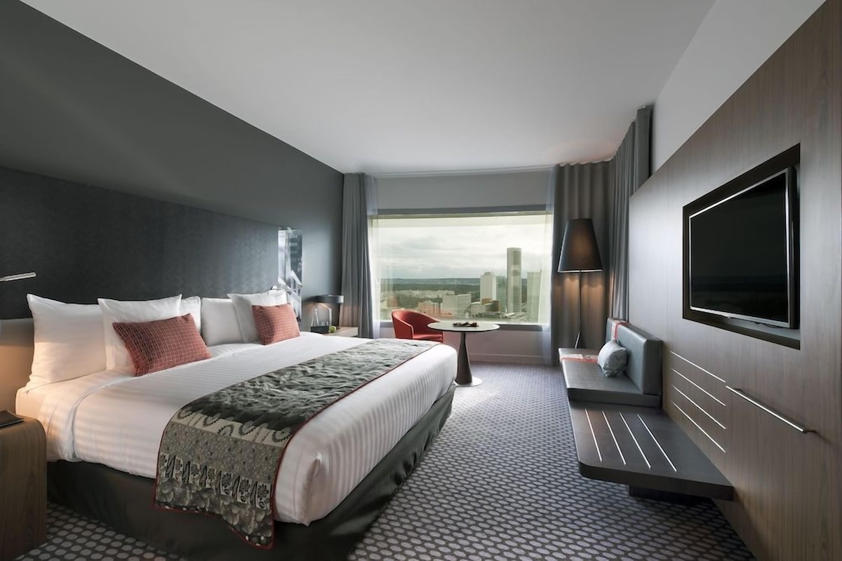 A modern hotel room with a large bed, a seating area, a flat-screen TV, and a city view.