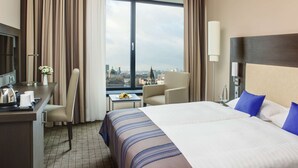 Habitación de hotel con una cama grande, un escritorio, una silla y vistas a un paisaje urbano a través de un ventanal.