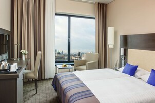 Ein Hotelzimmer mit einem großen Bett, einem Schreibtisch, einem Sessel und Blick auf eine Stadtkulisse durch ein großes Fenster.