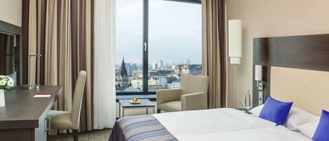 A hotel room with a large bed, a desk, a chair, a small table, and a view of a cityscape through a large window.