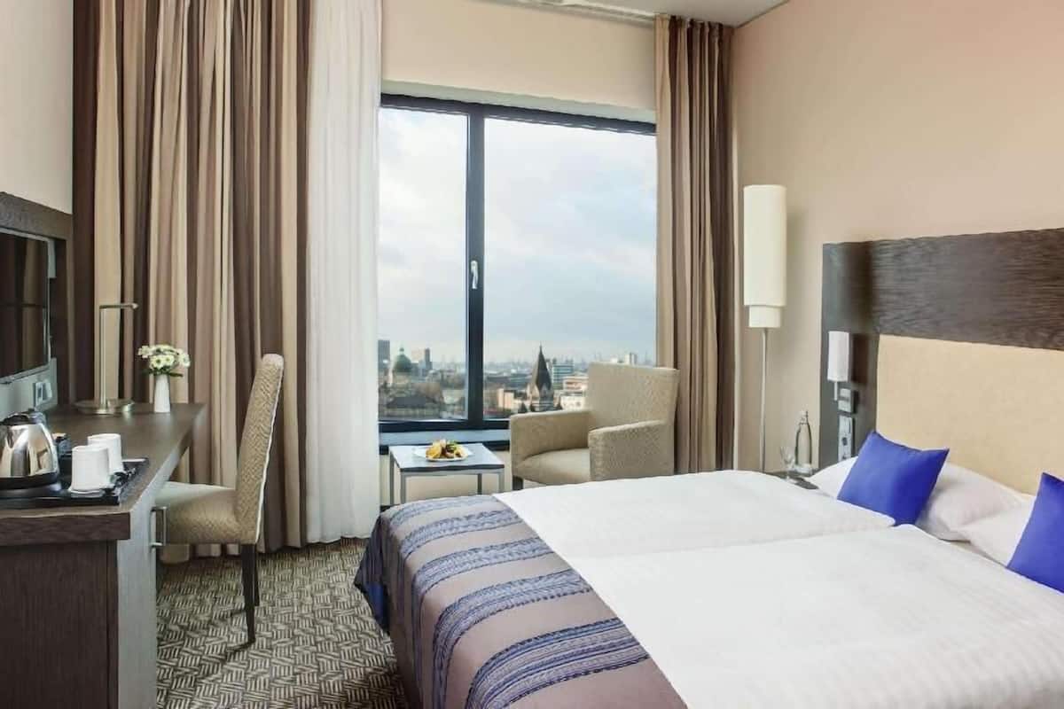 Une chambre d’hôtel avec un grand lit, un bureau avec une chaise, une petite table avec une lampe, et une vue sur la ville par une grande fenêtre.