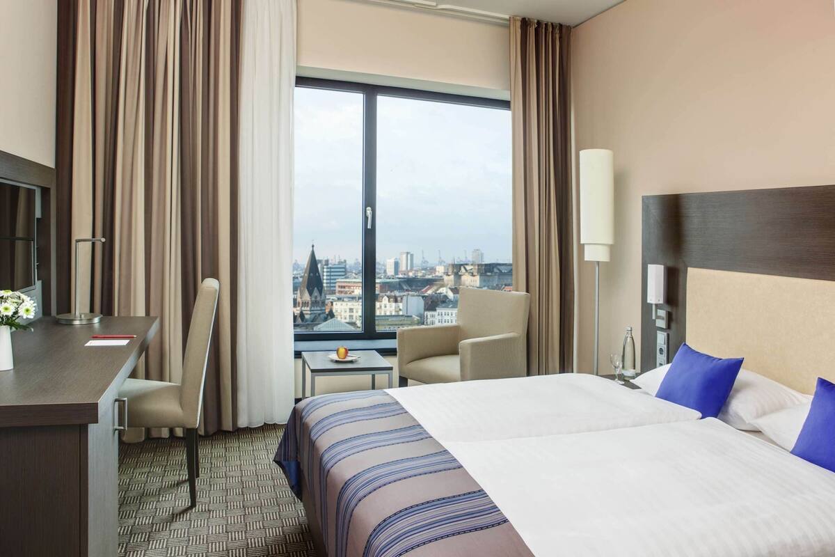 Une chambre d’hôtel avec un grand lit, un bureau, une chaise, une petite table et une vue sur le paysage urbain.