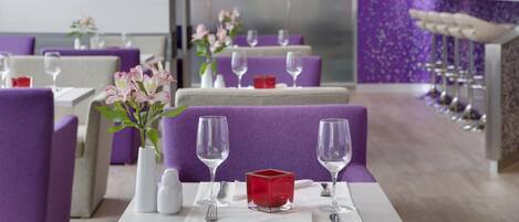 Un restaurante con sillas tapizadas de color morado, vajilla blanca y un centro de mesa en forma de cubo rojo.