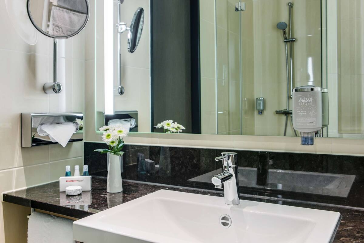 Une salle de bain moderne avec un lavabo blanc, un miroir, un distributeur de serviettes et un espace douche.