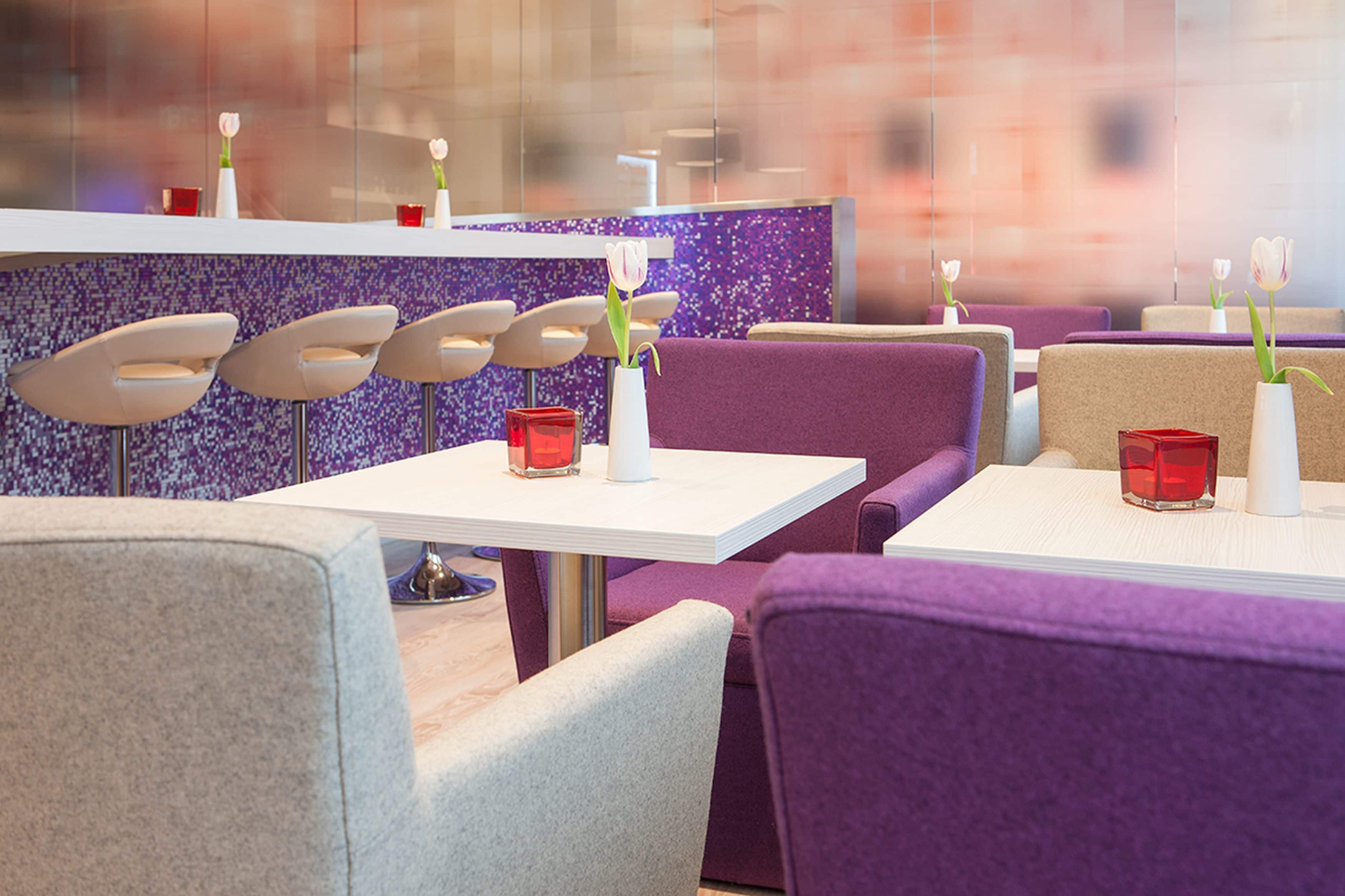 Ein Restaurant mit violetten und weißen Sitzgelegenheiten, roten Kerzenhaltern und weißen Vasen mit Blumen.
