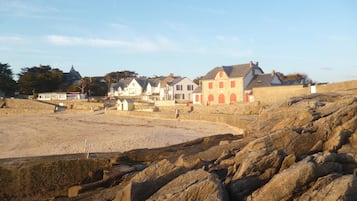 A coastal area with a rocky foreground, a sandy beach, and several buildings with distinct architectural styles, including a prominent building with red accents.