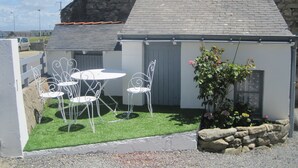 Un pequeño edificio con fachada de piedra, techo de tejas y un patio vallado con sillas y una mesa de metal blanco.