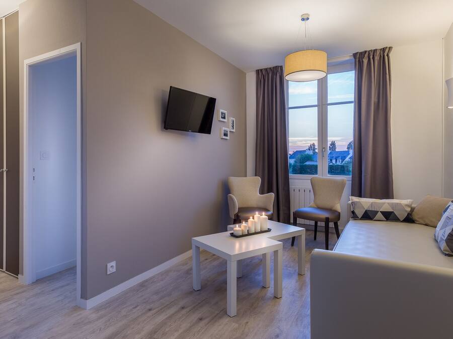 Un salón moderno con un sofá, un televisor montado en la pared, una mesa con velas y vistas a la ciudad a través de amplios ventanales.