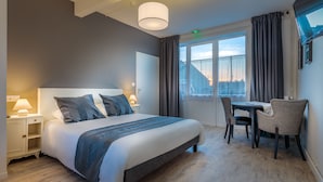 Habitación de hotel con cama, mesitas de noche, un escritorio con sillas, un ventanal con cortinas y una lámpara en el techo.