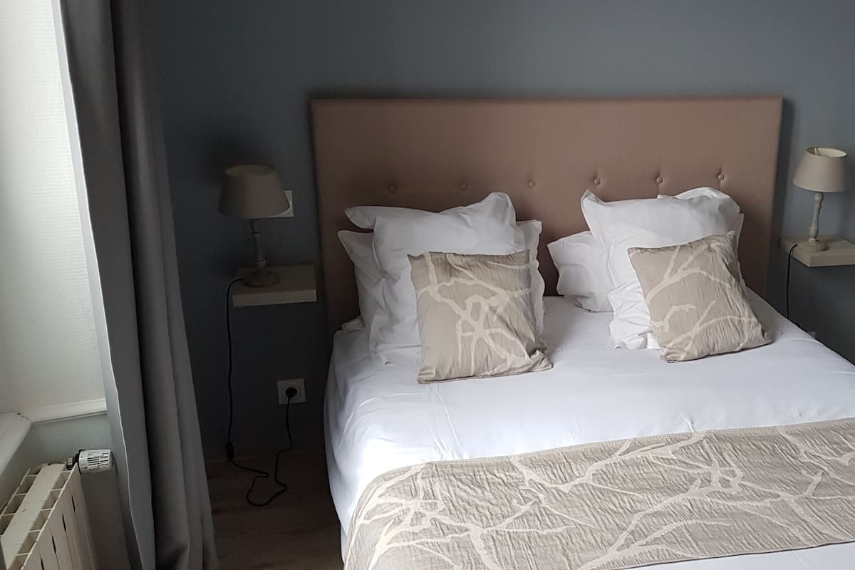 Una cama bien hecha con una colcha estampada y dos almohadones decorativos, una mesita de noche con una lámpara, y una puerta con interruptor de luz.