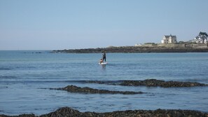 A coastal scene with a person on a paddleboard, calm water, rocky shoreline, and a castle-like building in the background. - Hôtel des Marais Salants (Batz-sur-Mer)