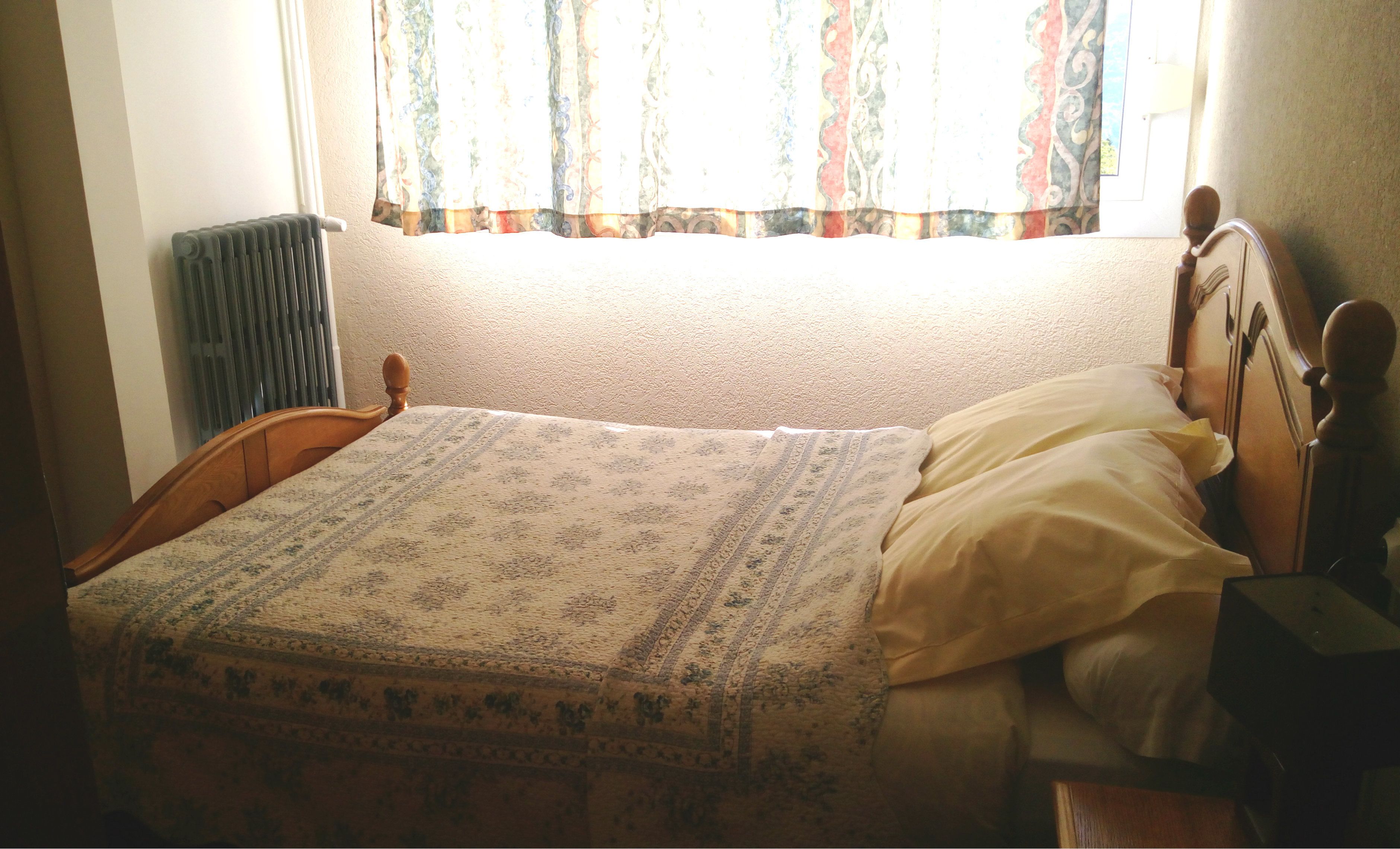 Een enkel bed met een bedovertrek met patroon en kussens, een radiator en een raam met gordijnen.