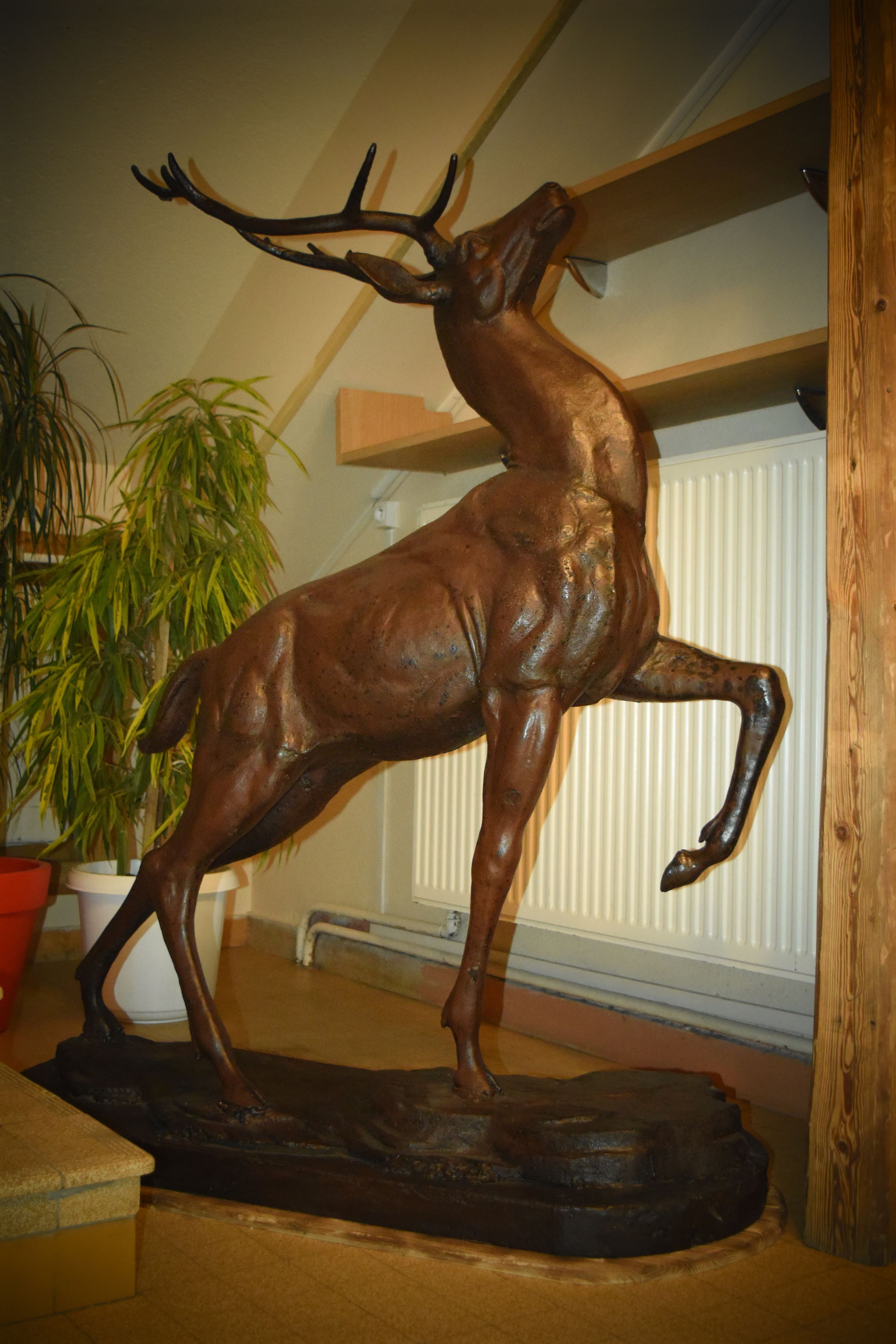 A bronze deer sculpture on a wooden base, positioned in a room with a potted plant and a radiator in the background.