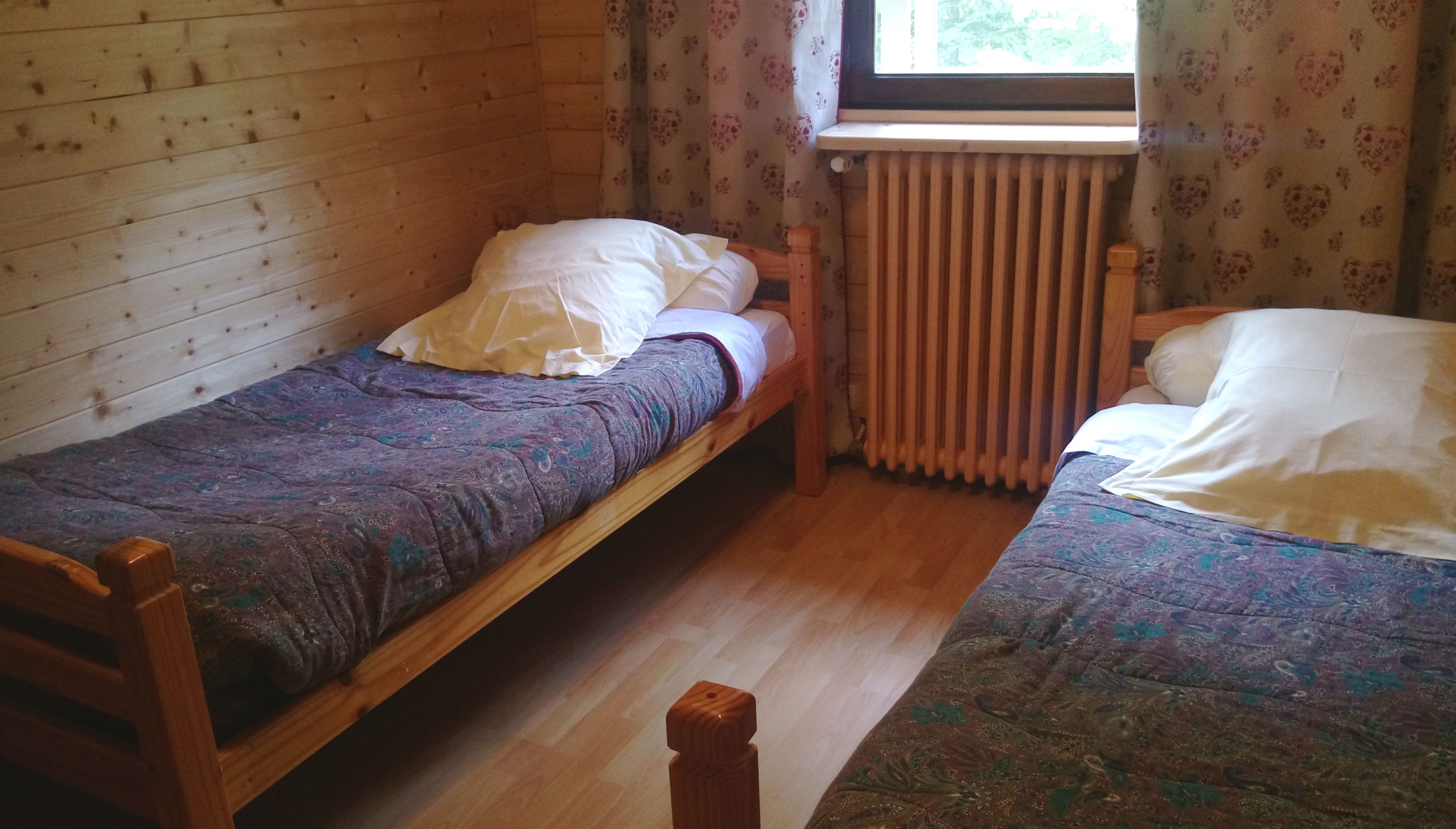 Une petite pièce chaleureuse, avec deux lits en bois, un radiateur et une fenêtre ornée de rideaux à motifs floraux.