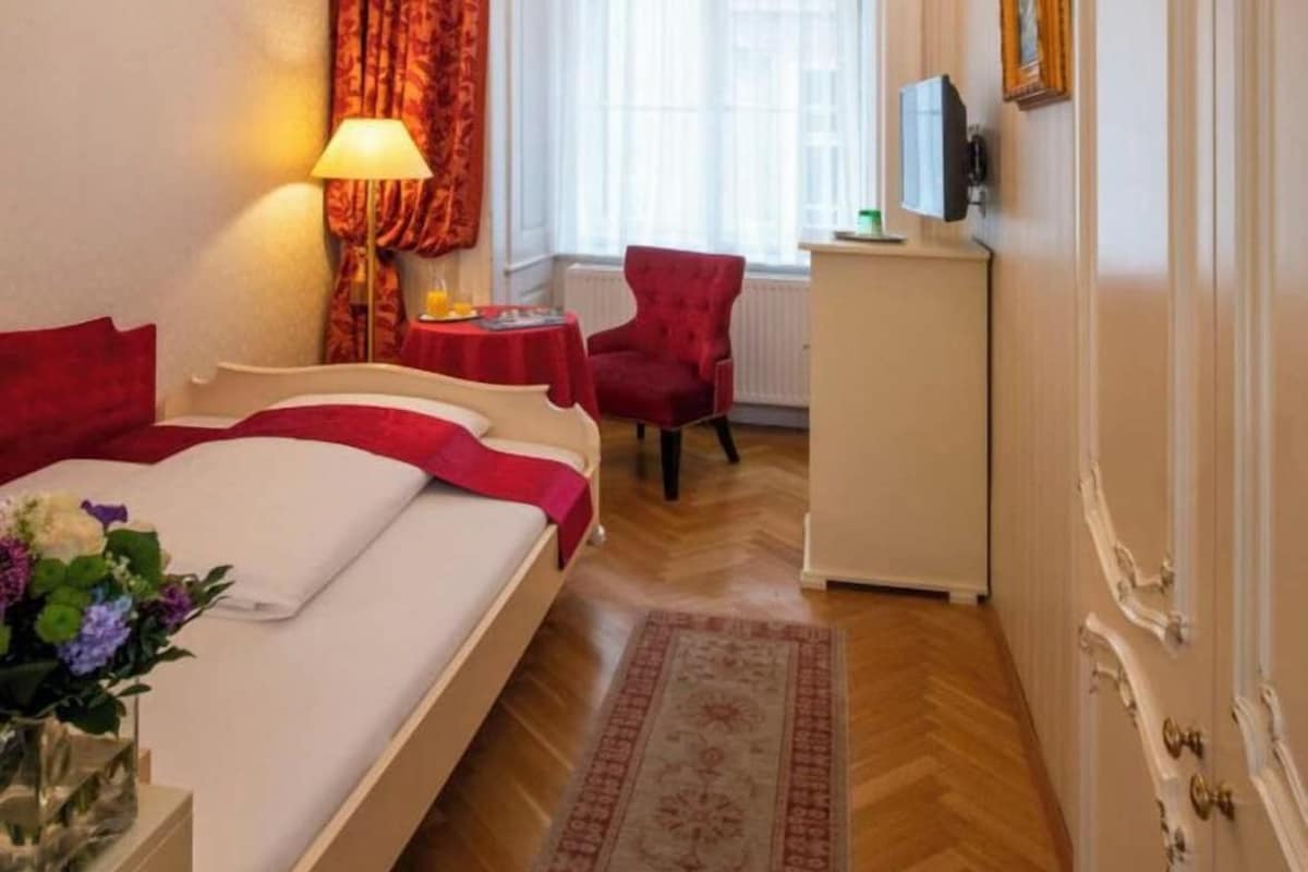 Habitación de hotel con cama, escritorio, silla, cortina roja, candelabro y vista al exterior.