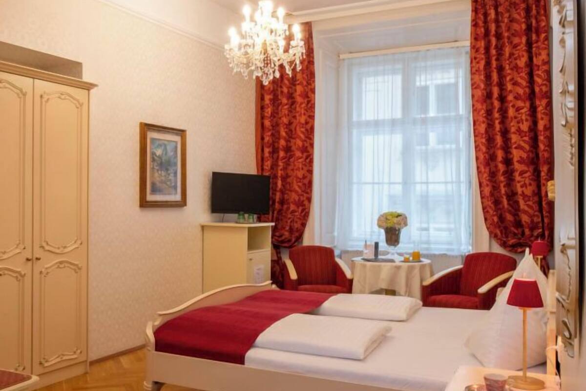 Um quarto com uma cama, um candelabro, uma televisão, uma mesa pequena e uma janela com cortinas.