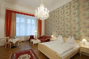 Een hotelkamer met een groot bed, een kroonluchter, een eettafel met stoelen, een rode fauteuil en behang met een patroon.