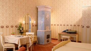 A hotel room with a bed, a small table with a vase of flowers, a chair, a desk with a suitcase, and a decorative pillar.