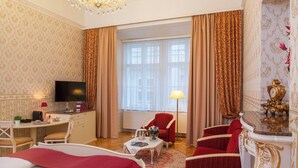 A luxurious hotel room with a large window, a chandelier, a desk with a television, and a red armchair.