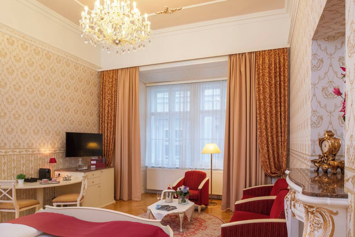 Quarto de hotel luxuoso com uma grande janela, um candelabro, uma escrivaninha com televisão e uma poltrona vermelha.