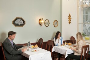 Et spiseområde med borde dækket til tre personer, hver med mad, glas og en flaske. Væggene er dekoreret, og et vindue giver udsigt.
