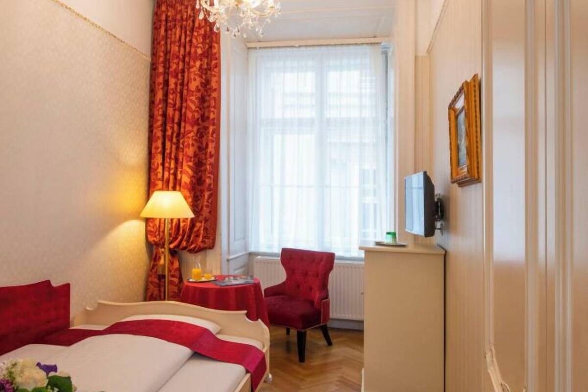 Quarto de hotel com cama, escrivaninha, cadeira, lustre e janela com cortinas.