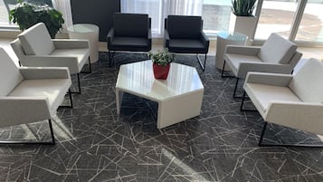 Een moderne zithoek met een glazen salontafel, omringd door vier grijze fauteuils en twee banken. Een potplant staat op de salontafel. De vloer heeft een donker, gescheurd patroon.