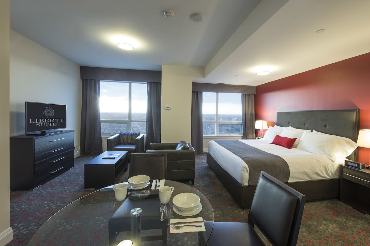 A hotel room with a bed, a TV, a dining area with a table set, and a view of the city through large windows.