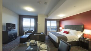 Habitación de hotel con cama, televisor, zona de comedor con mesa puesta y vista de la ciudad a través de amplias ventanas.