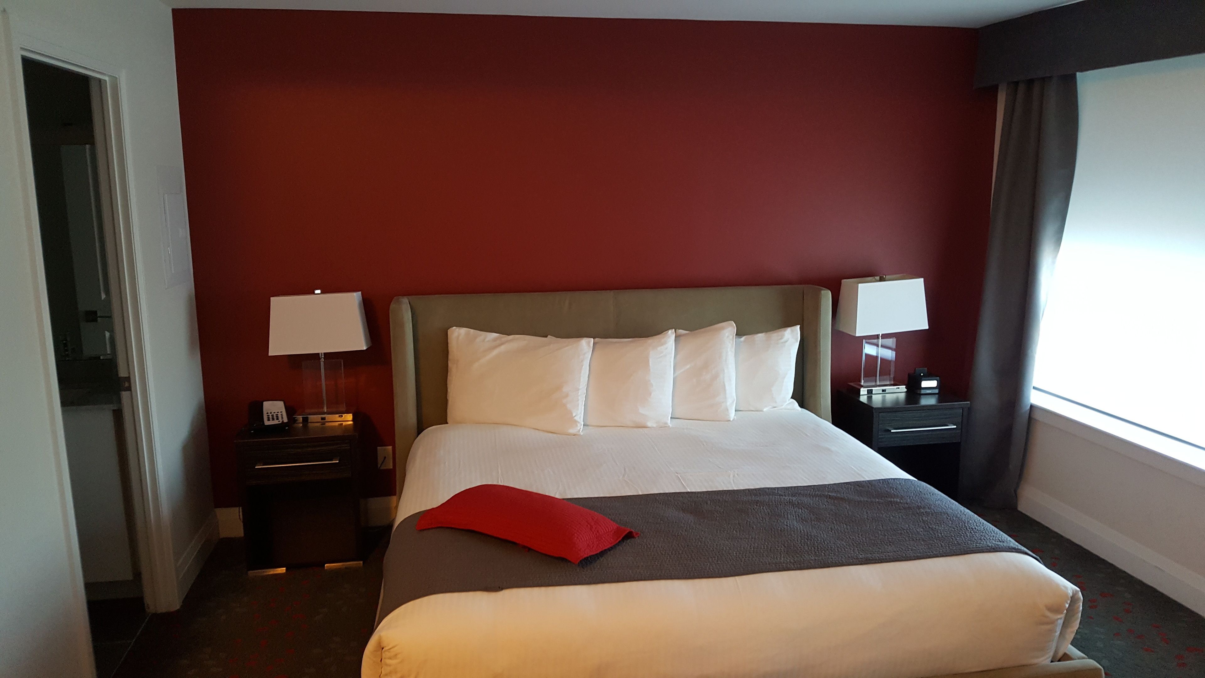 Una habitación de hotel con una cama grande, una mesita de noche con lámpara, un cojín rojo y una ventana con cortinas.