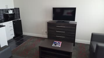 Un espacio habitable compacto con kitchenette, un televisor de pantalla plana sobre un mueble de madera y una revista sobre una mesa de centro.