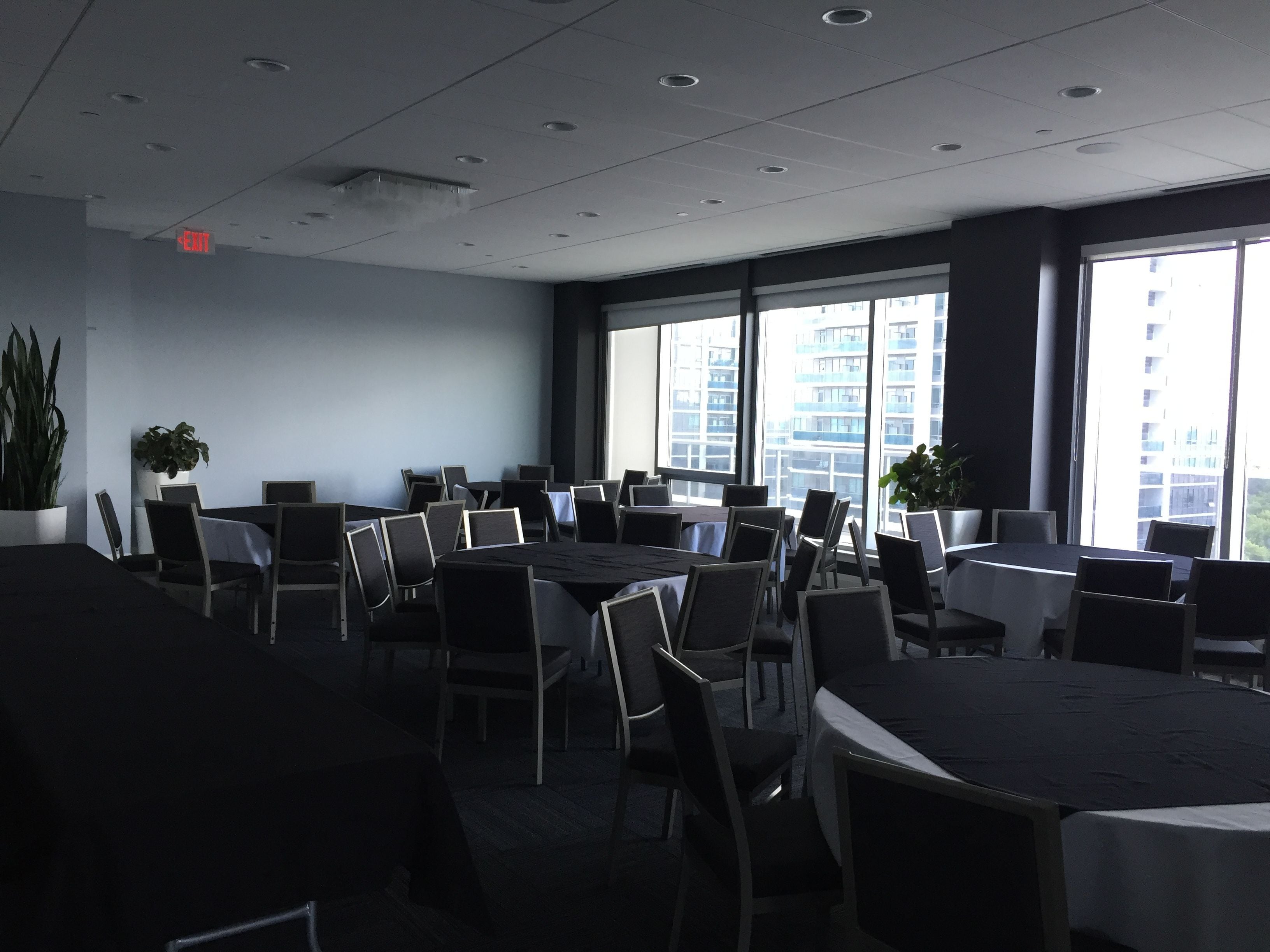 A conference room with multiple tables and chairs, large windows with a city view, and potted plants.