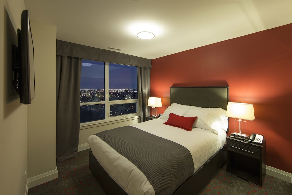 A hotel room with a large bed, two bedside tables with lamps, a TV mounted on the wall, and a window with a city view.