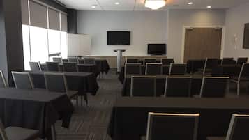 Une salle de conférence avec des rangées de tables et de chaises, un téléviseur accroché au mur et une porte au fond.