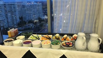 Un bufé con diversos alimentos y aderezos, una jarras y un bol de vegetales, con vista a la ciudad.