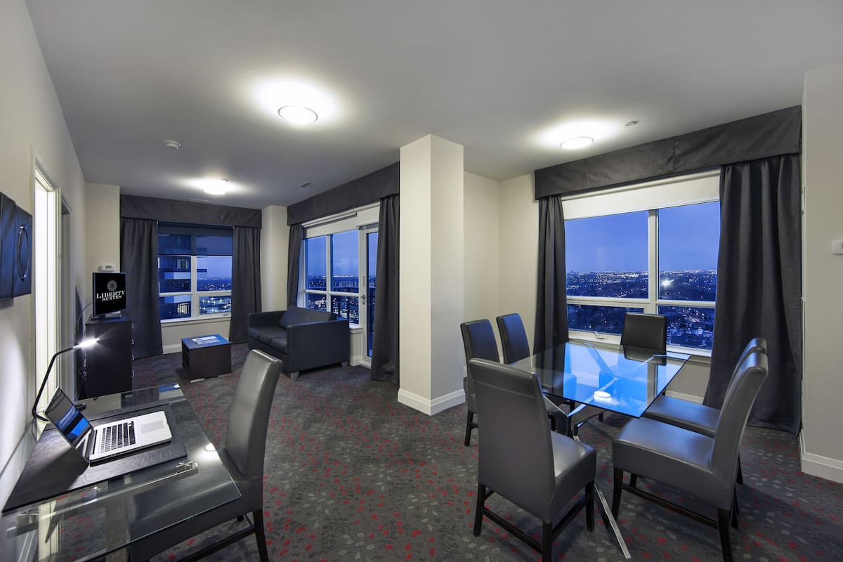 A modern hotel room with a glass table, leather chairs, a laptop, and a large window offering a city view.