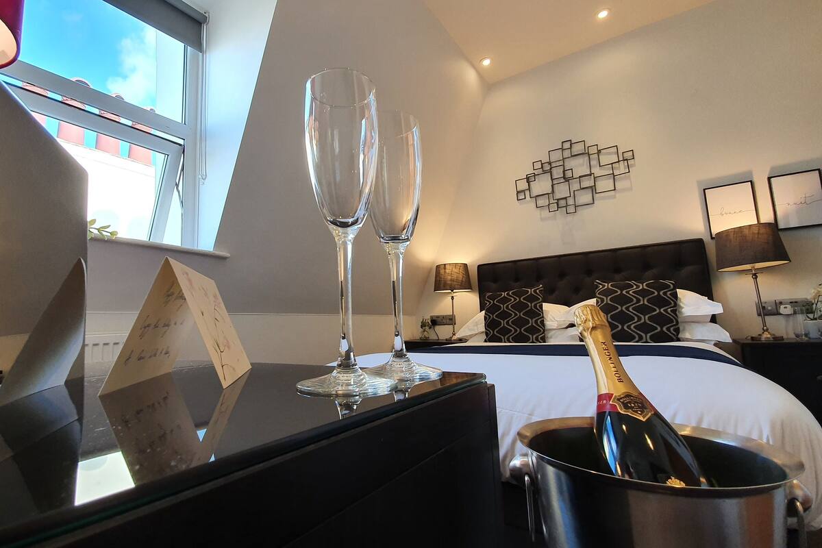A hotel room with a bed, bedside tables, a champagne bottle, and two champagne flutes on a table.