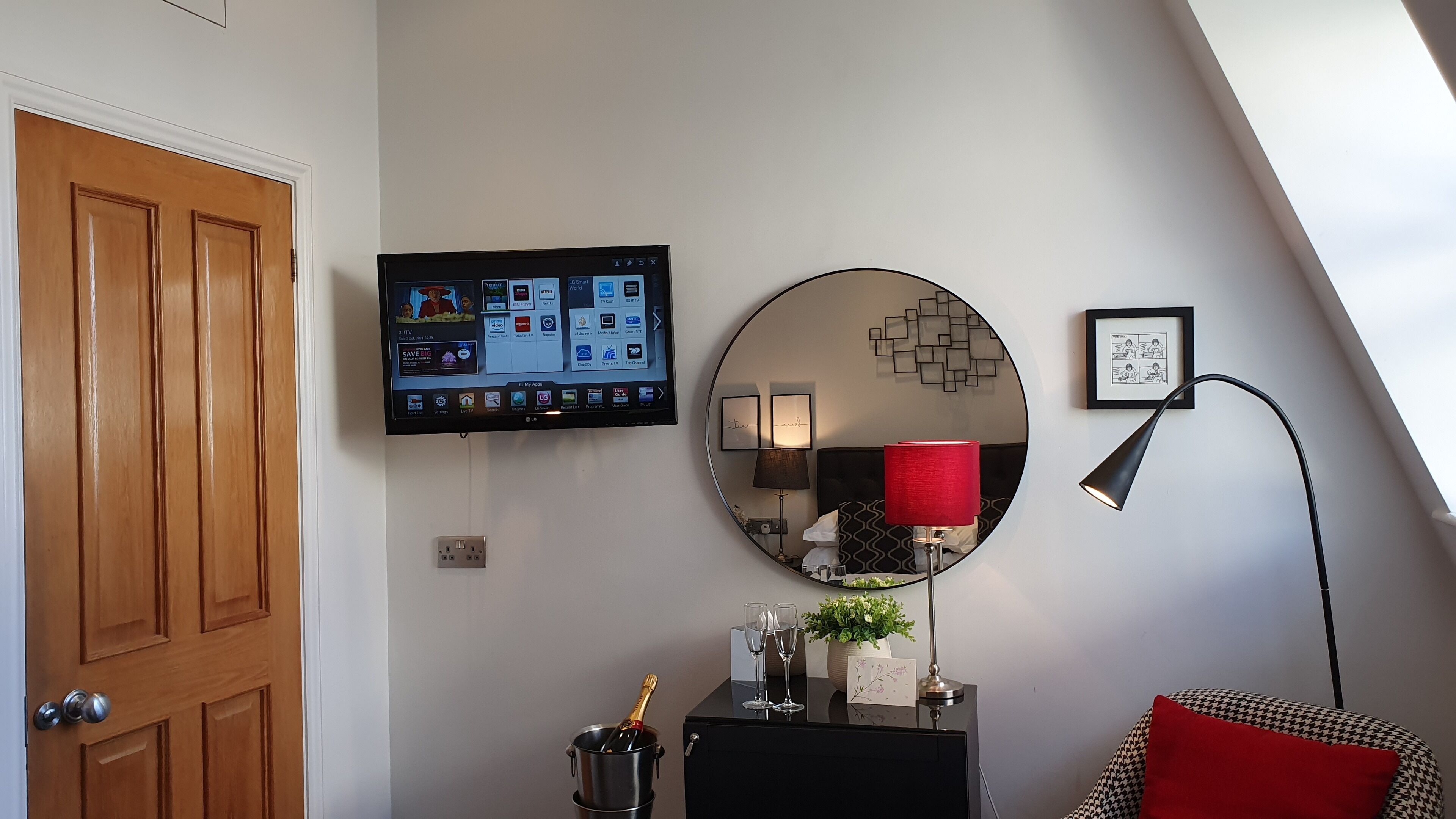 Ein modernes Wohnzimmer mit einem Wand-Flachbildfernseher, einem runden Spiegel, einer roten Lampe und einem kleinen Tisch mit einer Champagnerflasche und Gläsern.