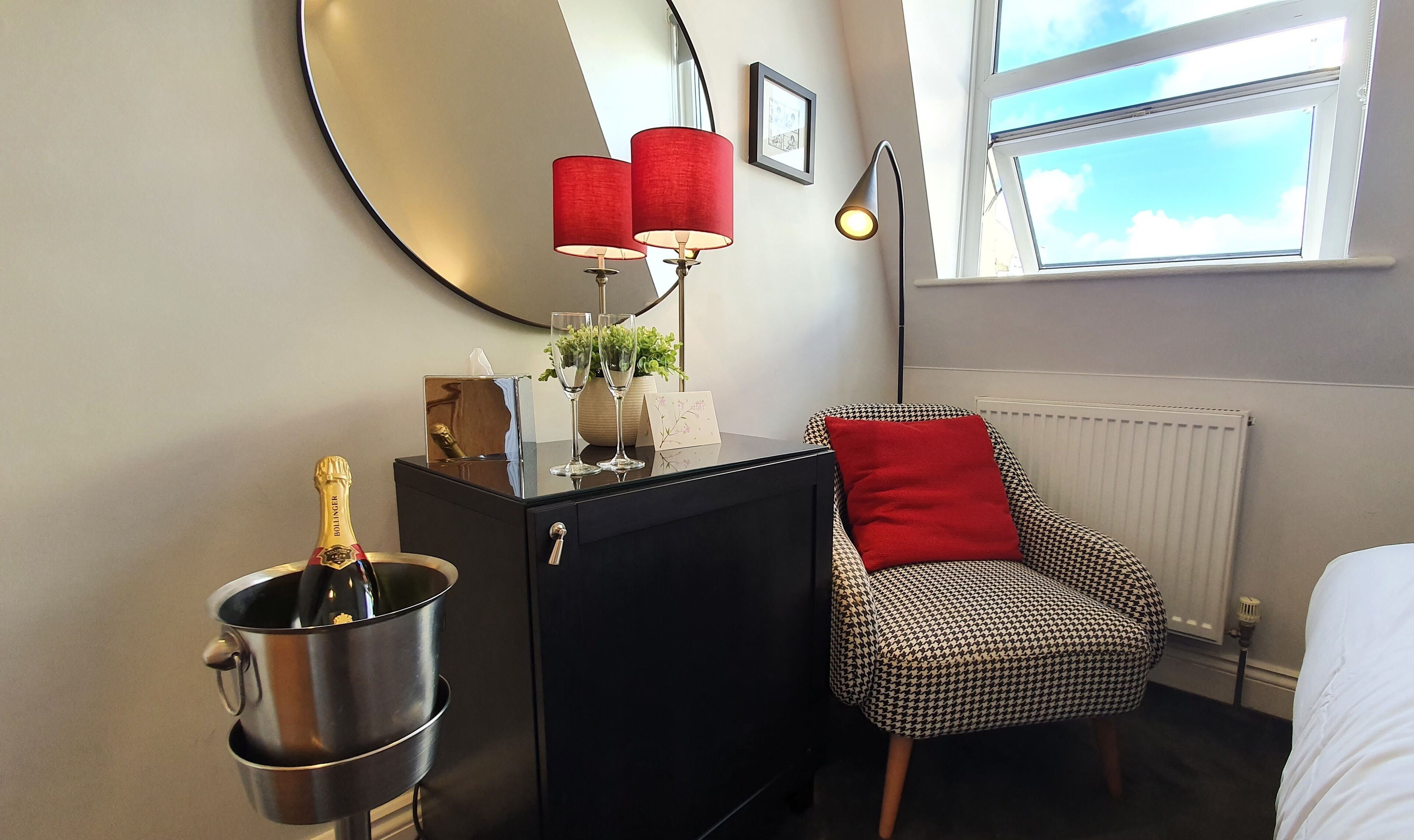 A hotel room with a bed, a desk with a computer, a chair, a red lamp, a window with a view of the sky, and a bottle of champagne.