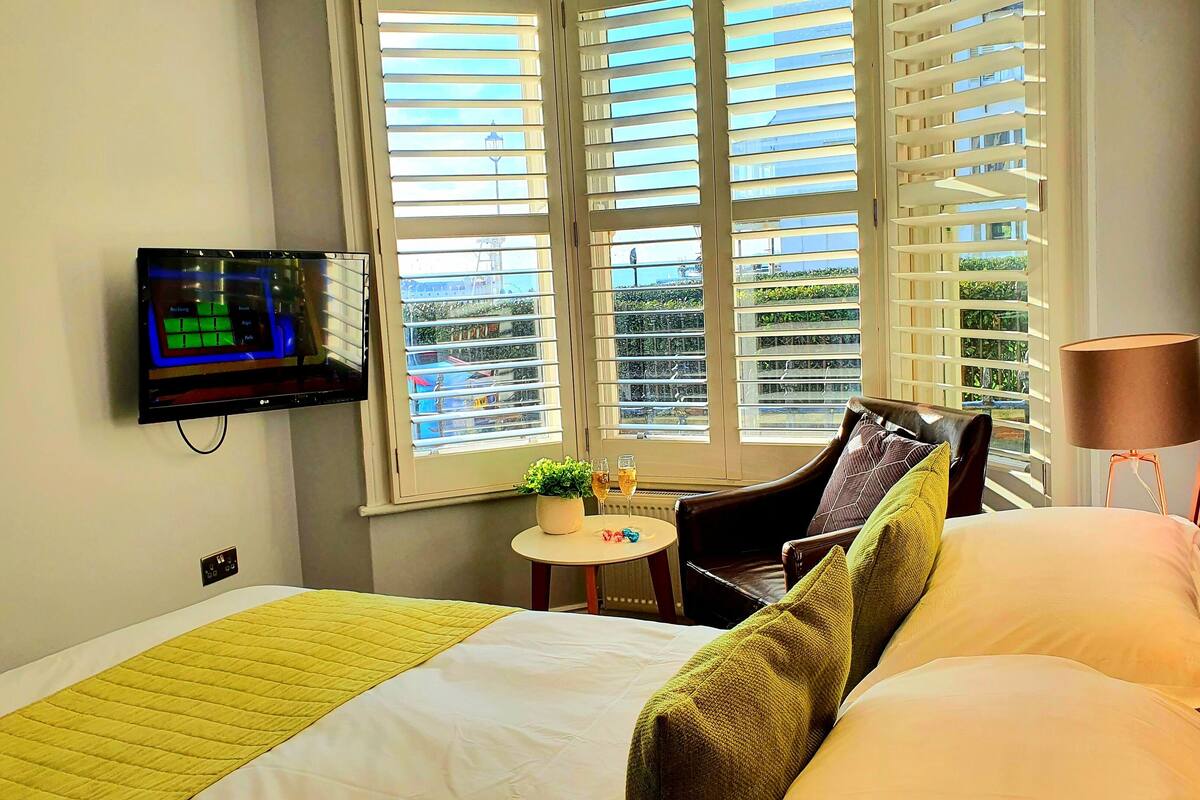 A hotel room with a bed, a chair, a small table with drinks, and a TV mounted on the wall.