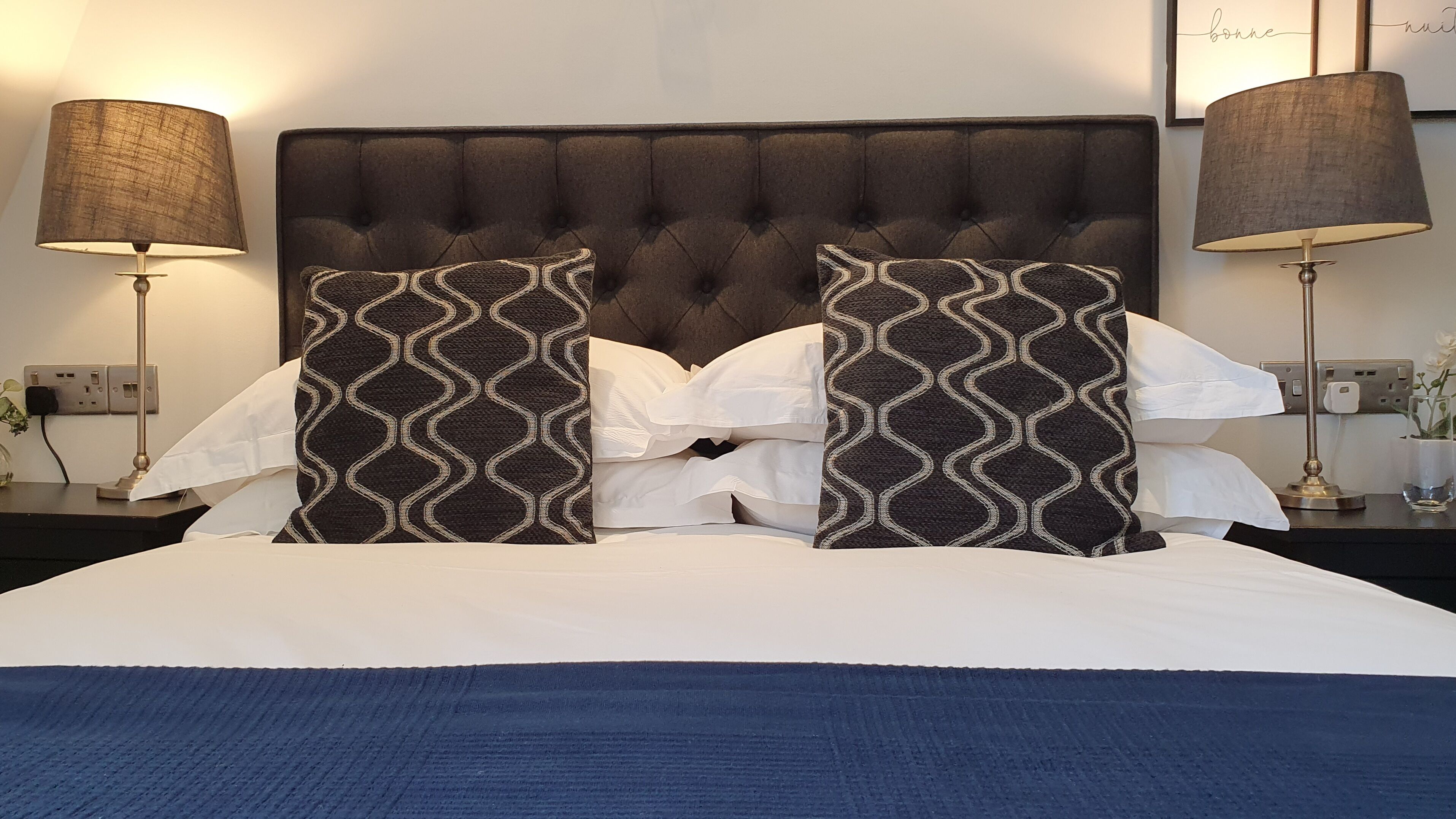 A neatly made bed with a tufted headboard, two patterned pillows, and two bedside lamps on a nightstand.