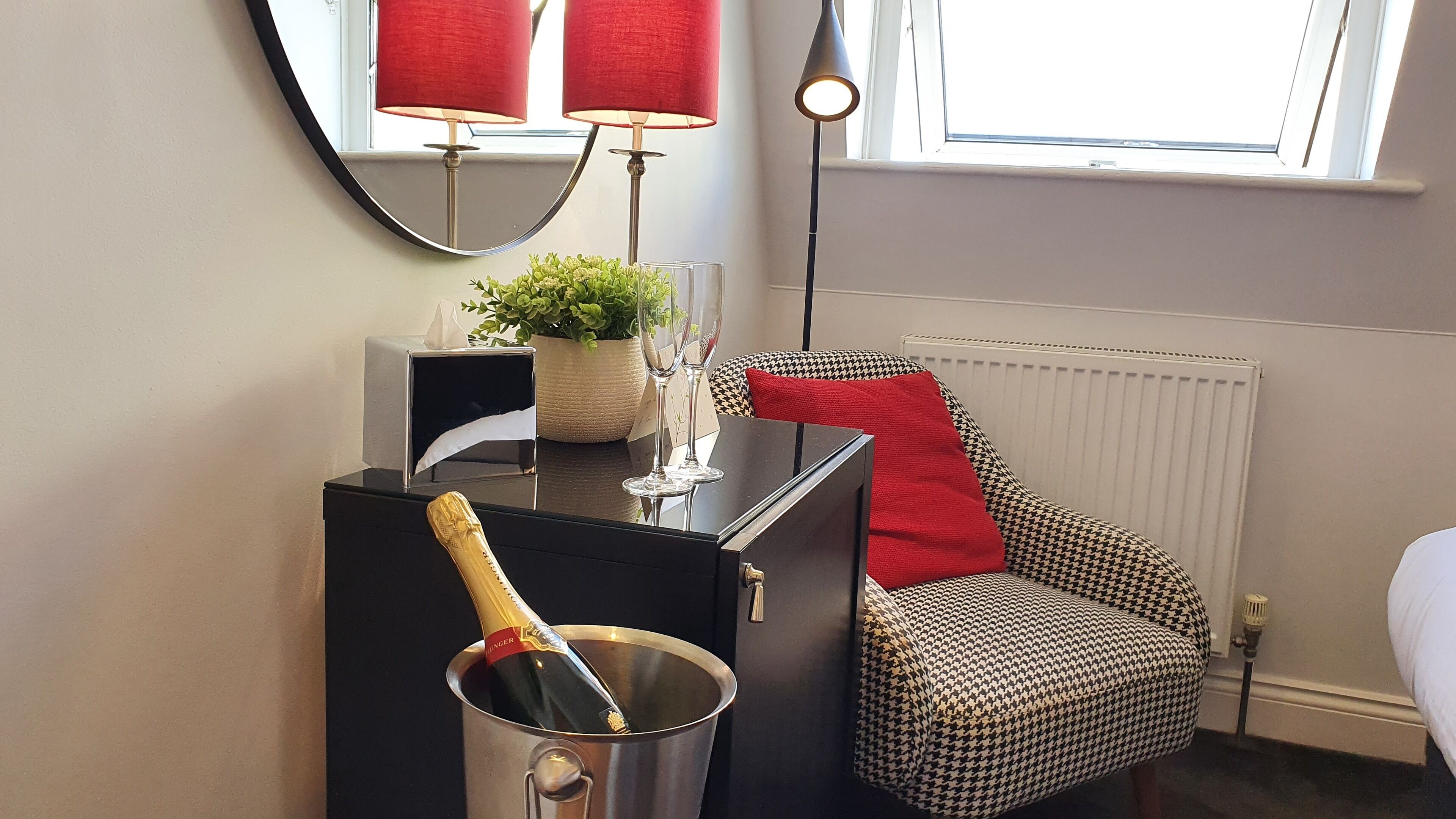 Una habitación con minibars negros, un cojín rojo en una silla de pata de gallo, una planta en maceta y un espejo con lámparas rojas.